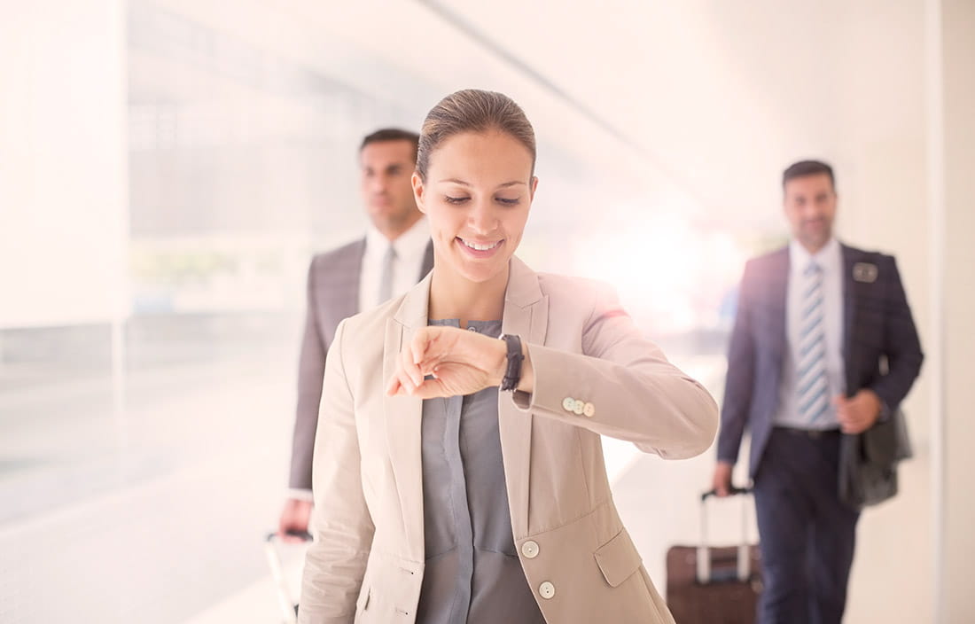 Smiling woman looking at her watch, walking through an airport with 2 people behind her.