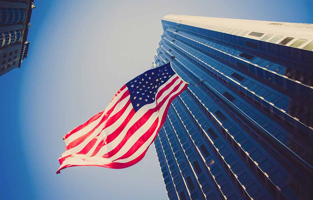 Picture looking up at an American flag waving in front of a skyscraper.