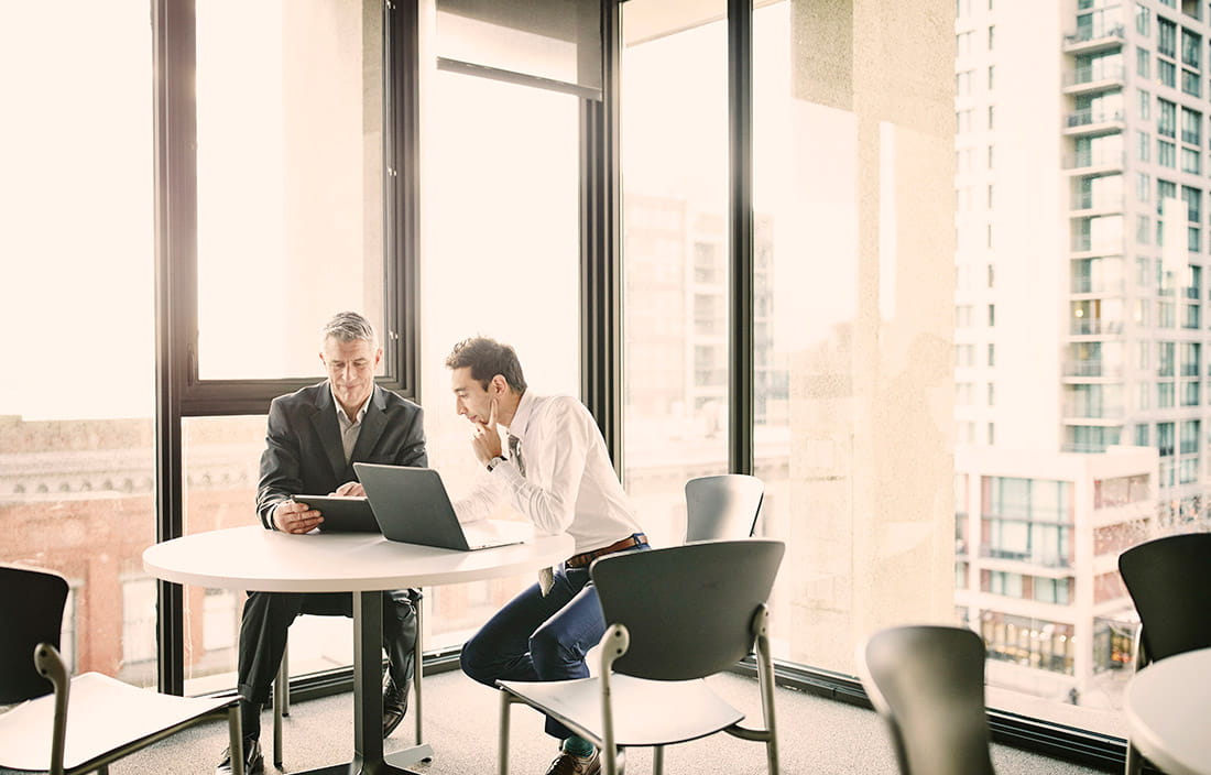 Two men sitting at a table in front of windows looking at a computer