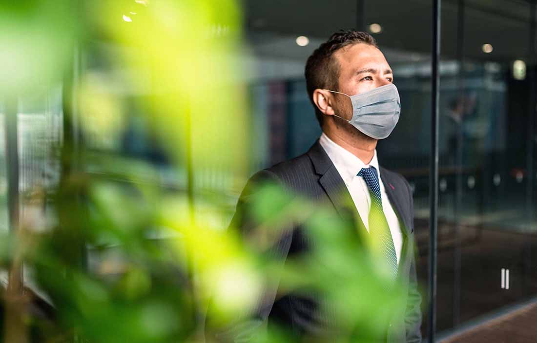 Man in a suit standing by a window wearing a mask