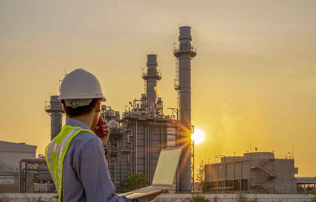View of a manufacturing worker standing holding a laptop next to a factory in the background.