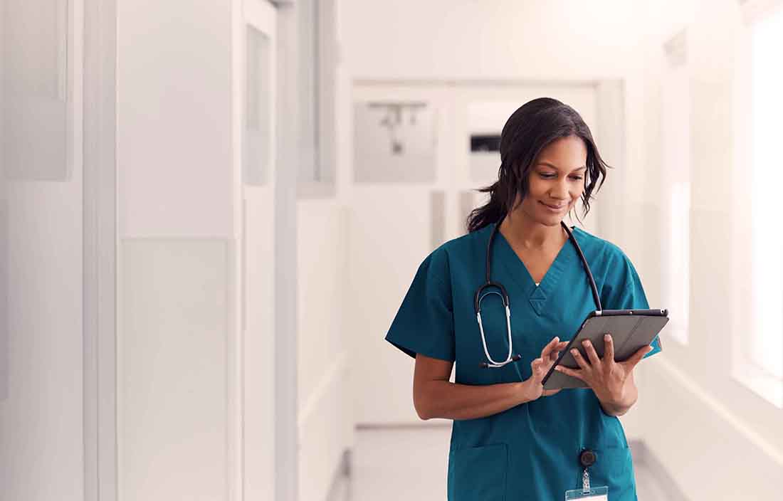Nurse smiling and writing notes in hospital hallway.