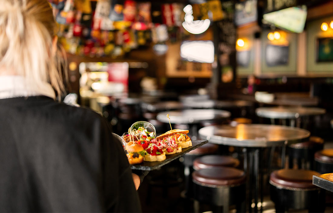 Server carrying a tray of appetizers in a sports bar, as seen from over her shoulder and with a blurry background.