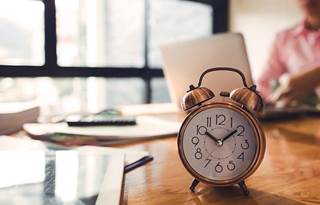 Clock on a desk next to a man on his laptop. 