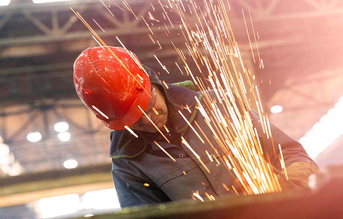Image of a man at a construction site wearing a red hardhat and safety glasses with sparks flying in front of him.