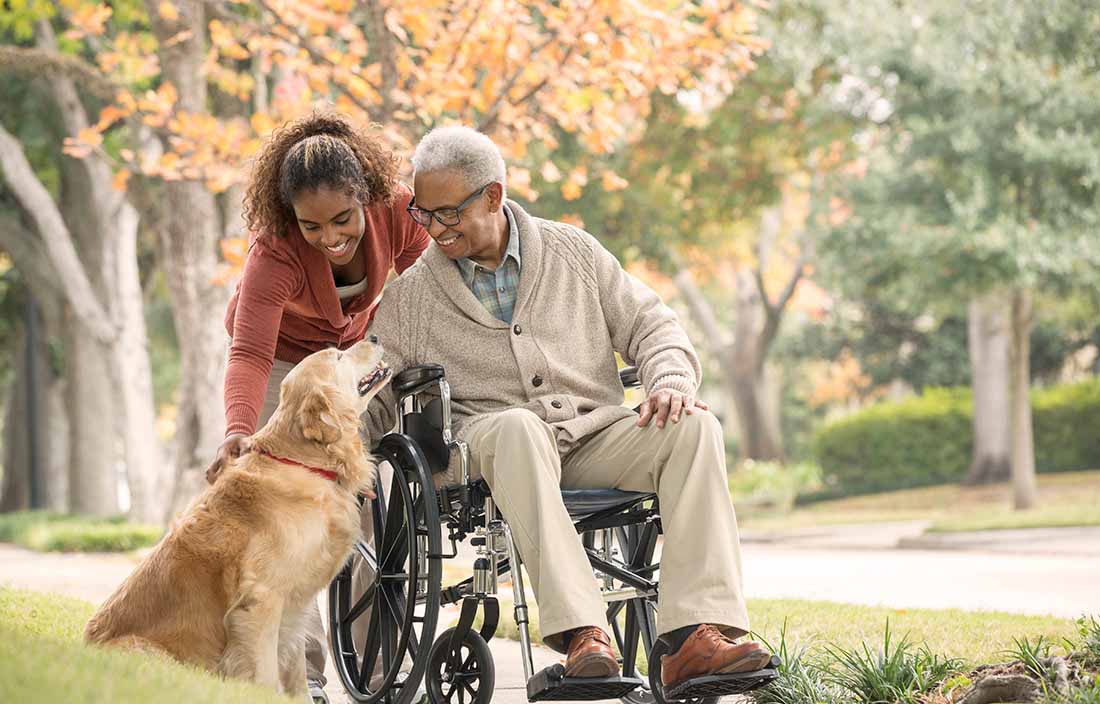 Image of a woman pushing an elderly man in a wheelchair outside and both petting a golden retriver dog.
