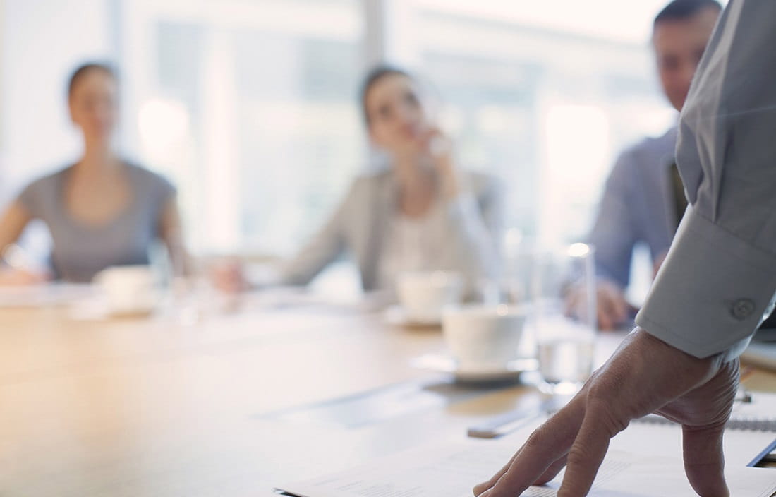 Image of businessman's hand on conference room table with other business people out of focus in the background.