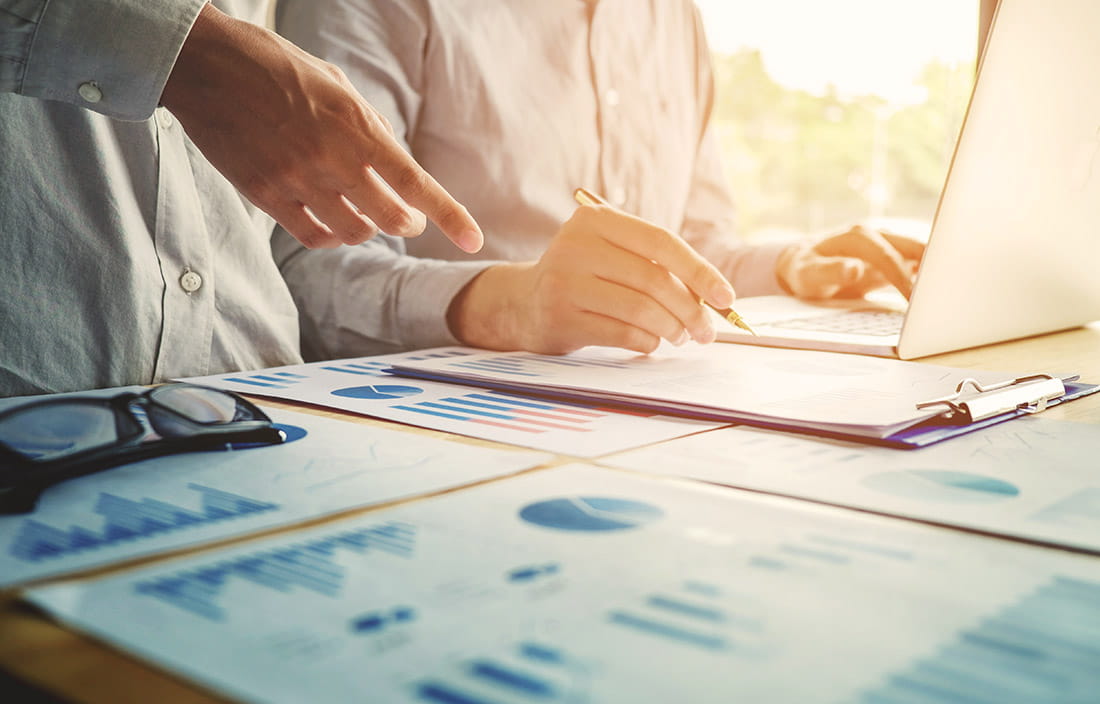 Image of desk with charts and business people working on a laptop.