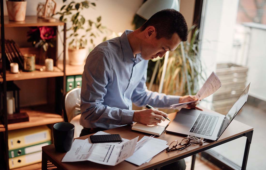 Man sitting at desk writing in a notebook with his laptop open