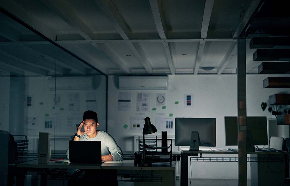 Man sitting at his desk looking at his computer monitor in an abandoned office at night.