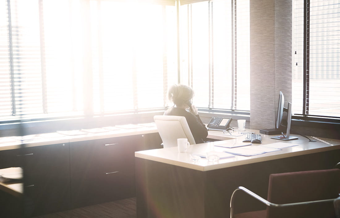A businesswoman sitting at her desk looking out an office window flooded with sunlight. 
