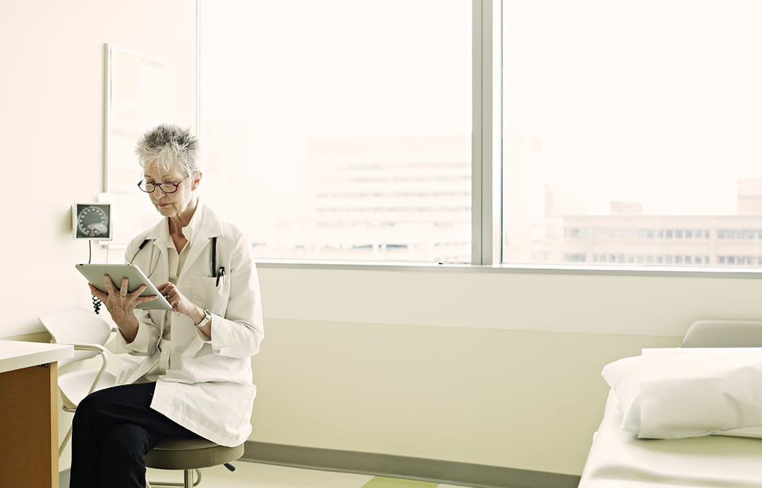 Physician in a treatment room sitting on a stool browsing on her tablet device. 