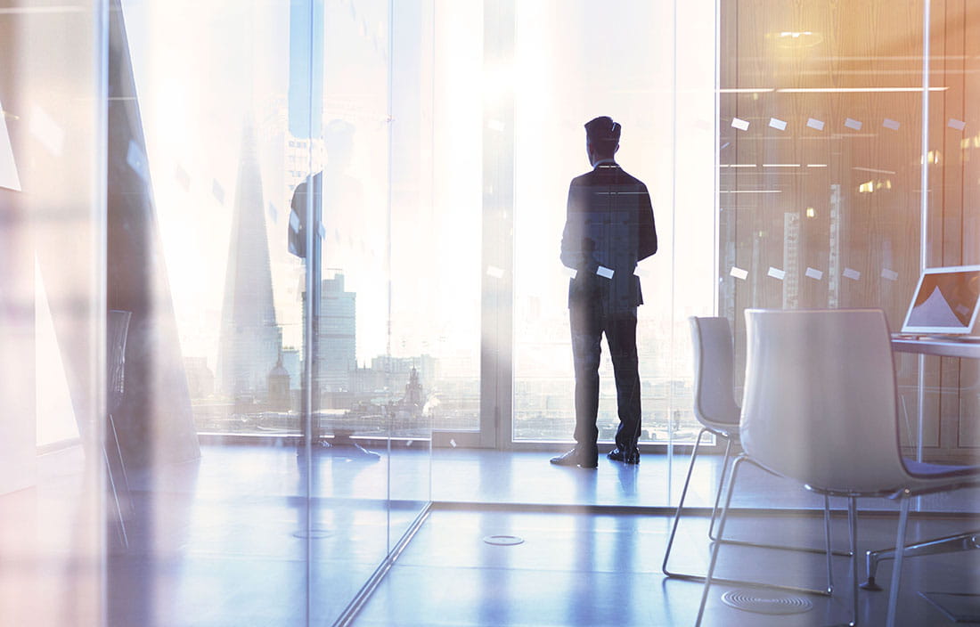 Businessman standing by a glass window in a corner office looking out the window at a city skyline. 