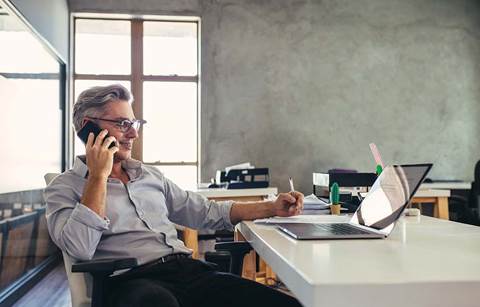 Man using his cell phone while writing with a pen on documents in front of him.