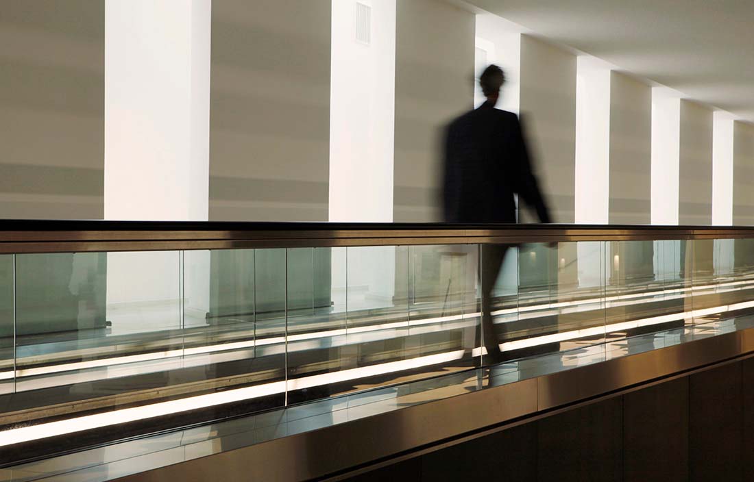 Picture of a blurry man walking down a corridor with glass railing to his right. 