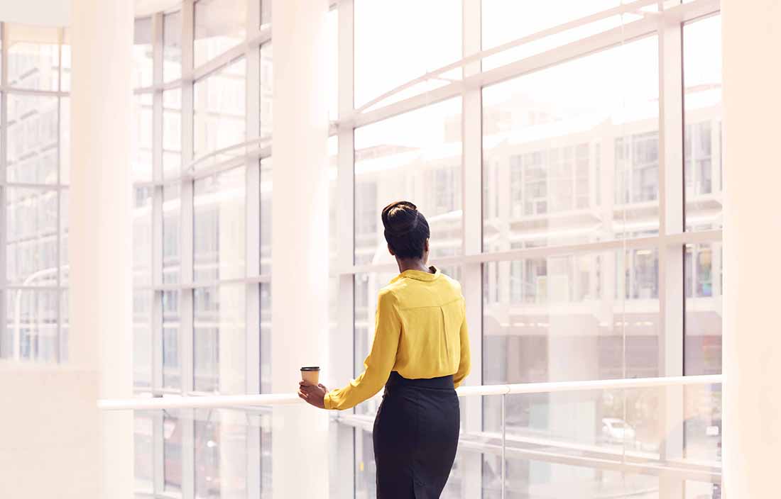 Business woman wearing yellow dress shirt standing in the lobby area of a building looking out the window.