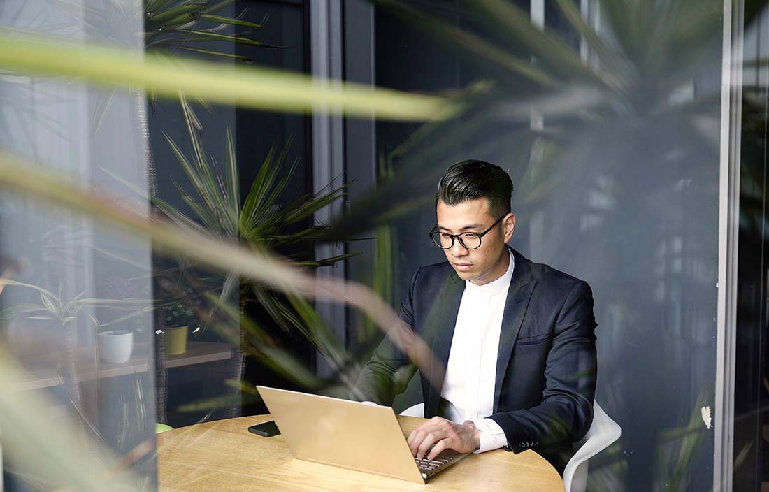 Young businessman sitting at a table reading about the economic impact on oil and gas companies on his laptop computer.