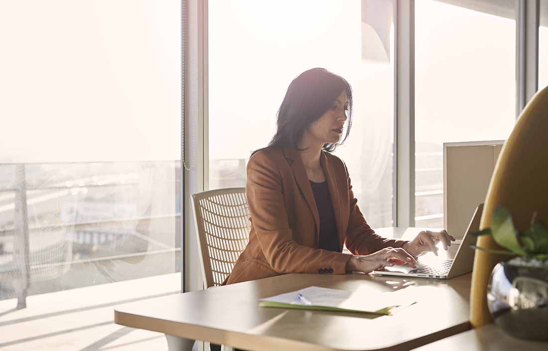 Businesswoman in a brown jacket sitting at a desk using a laptop computer.