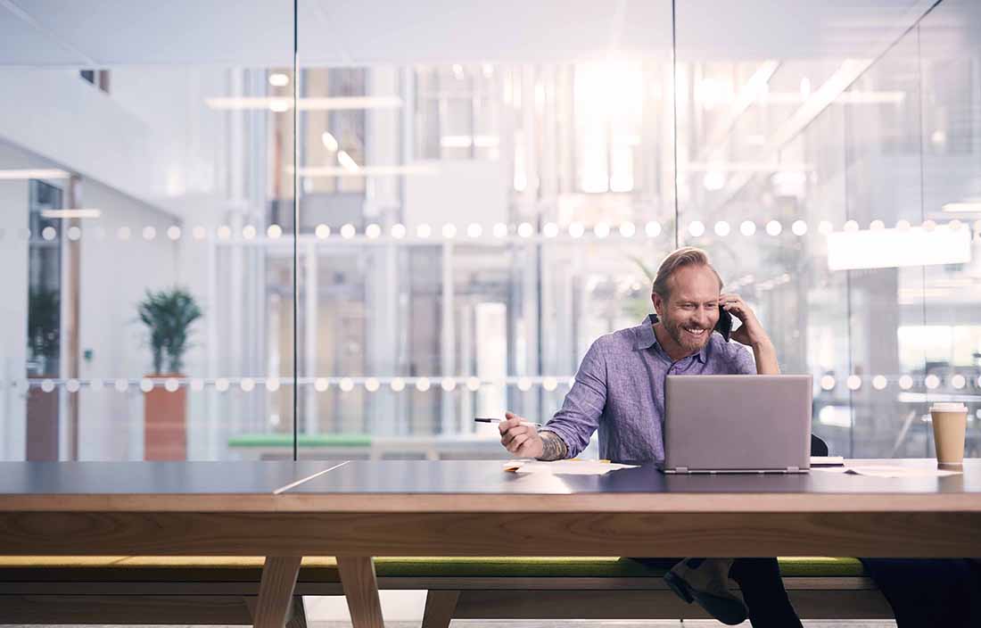 Man using cell phone smiling while sitting at a table using his laptop computer.