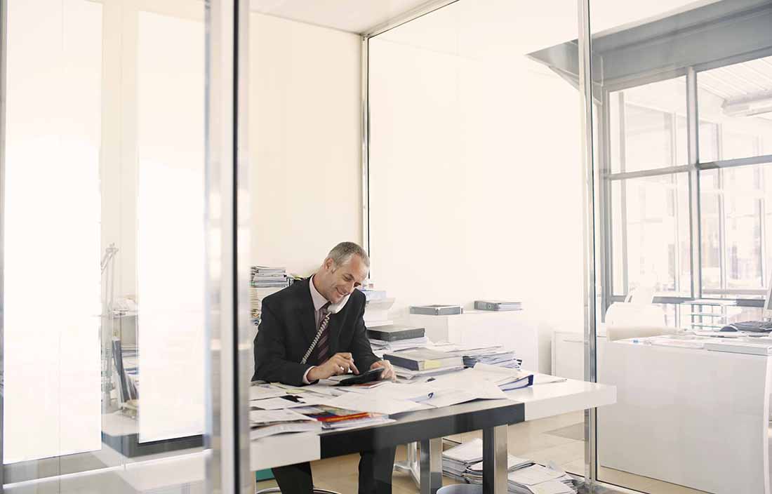 Man sitting at a messy desk, talking on the phone and typing on a calculator