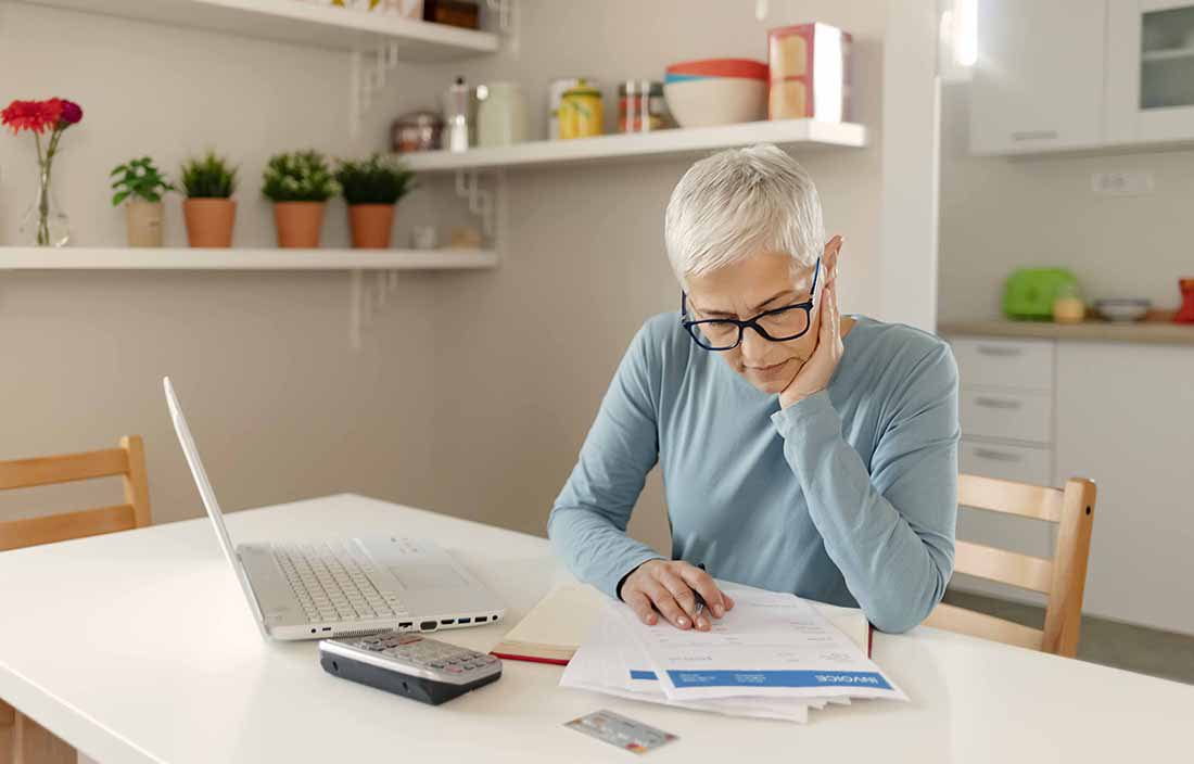 Elderly woman reviewing documents on a desk next to her laptop computer.