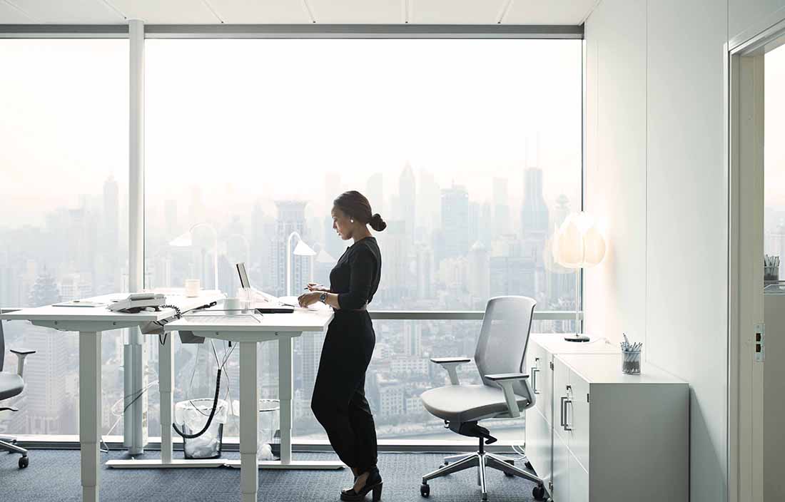 Woman in a glass office standing at a desk with a laptop