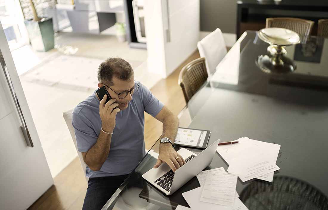 Man sitting at a table talking on his cellphone and typing on a laptop