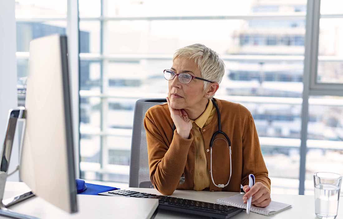 Elderly woman wearing a stethoscope while using a desktop computer.