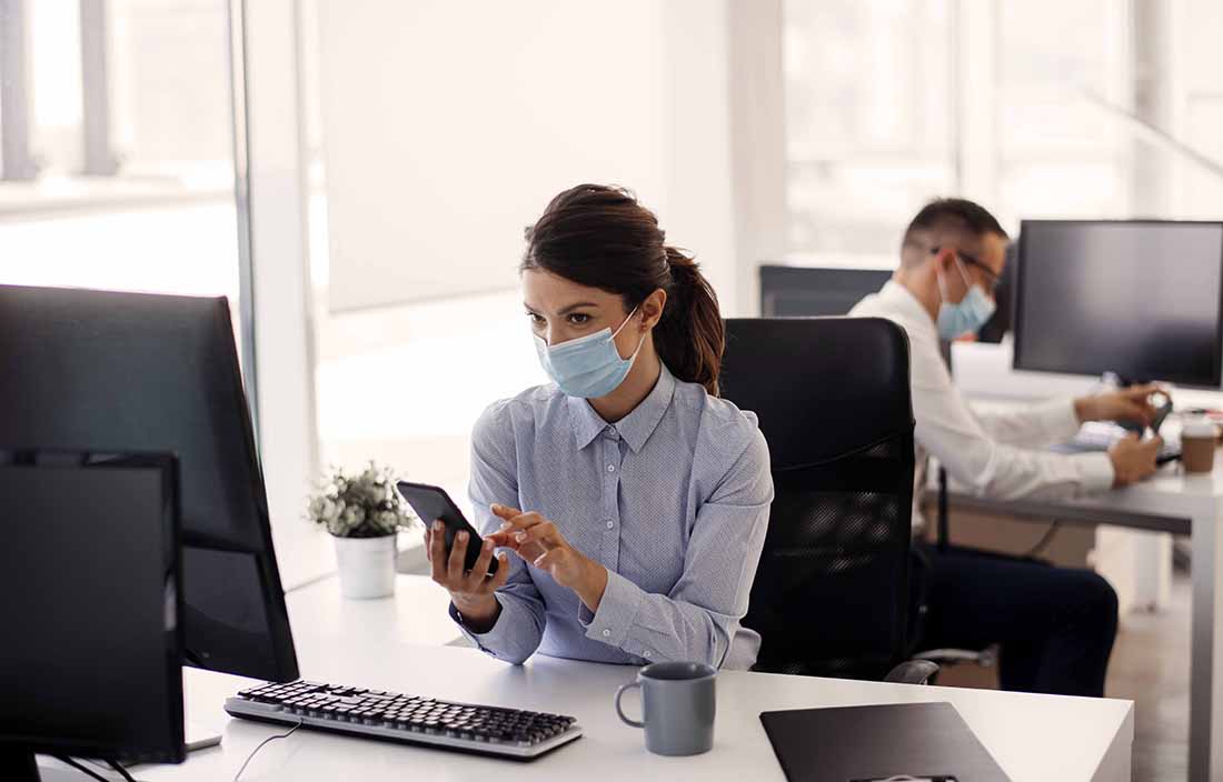 Woman wearing a mask while sitting at her desk, typing on her cellphone