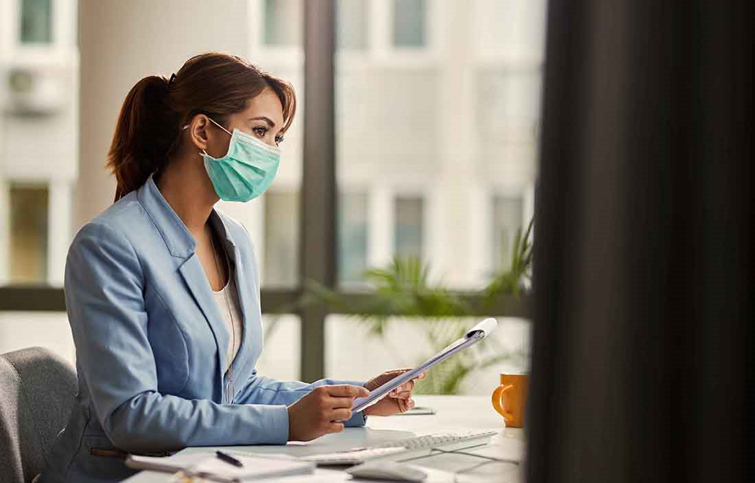 Businesswoman sitting at desk wearing a protective facemask while reviewing a notepad.