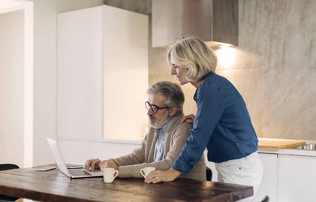 Elderly couple in the kitchen using a laptop computer while drinking coffee.
