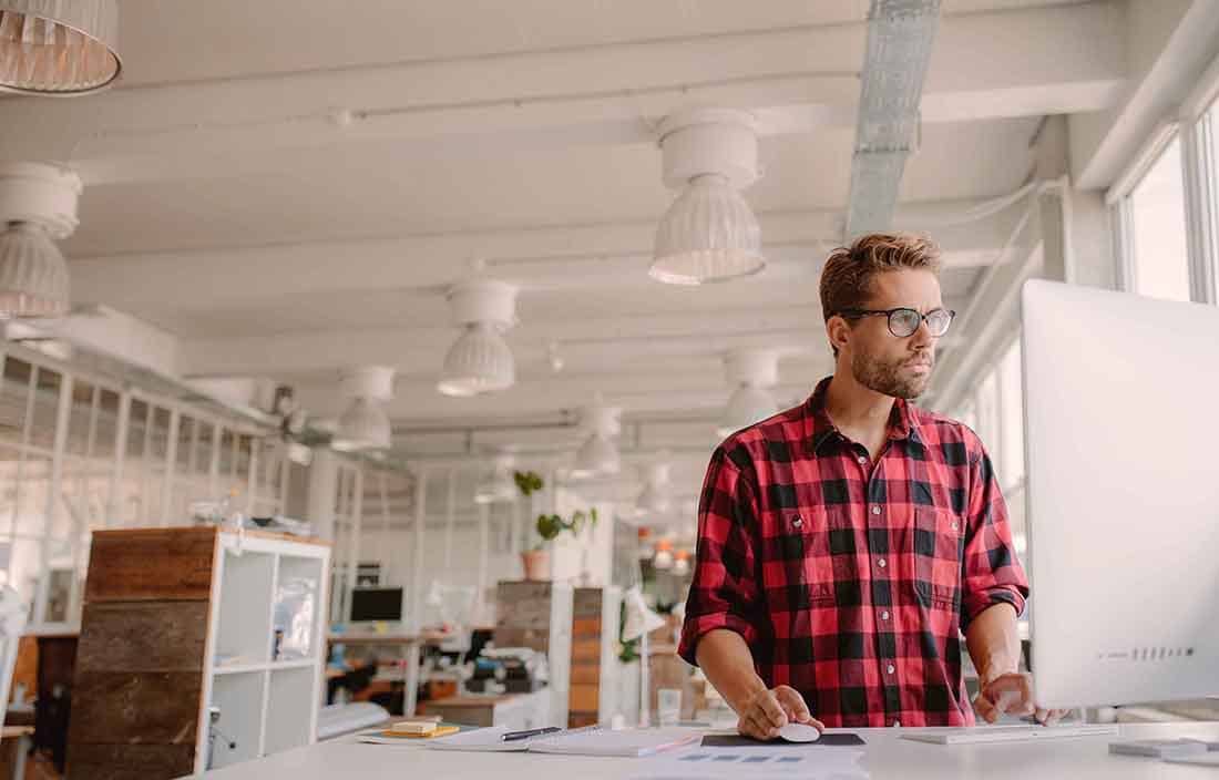 Man wearing casual clothes standing at a desk using a desktop computer.