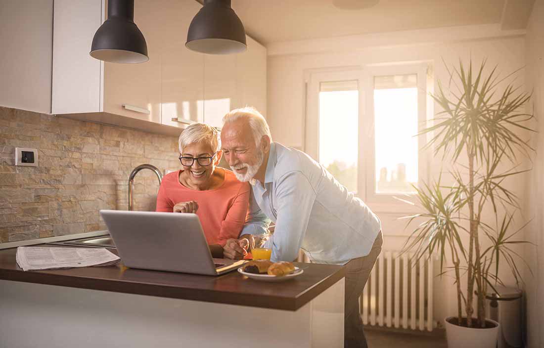An elderly couple in their kitchen using a laptop computer on the counter.