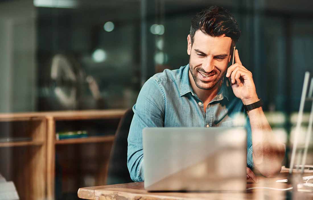 Man in a coffee shop taking a phone call while using his laptop computer.