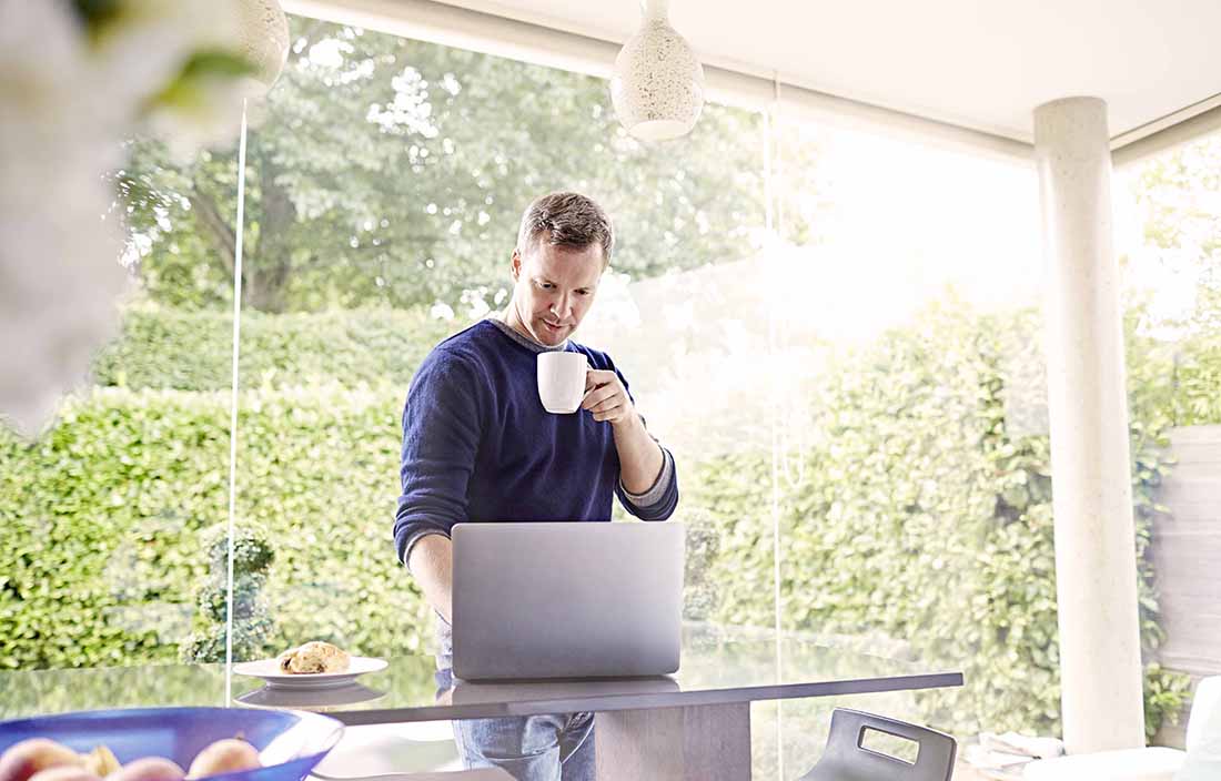 Man using laptop computer in kitchen while standing drinking a cup of coffee.