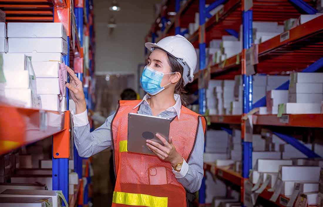 Woman in a warehouse wearing a hardhat checking inventory on warehouse shelving.