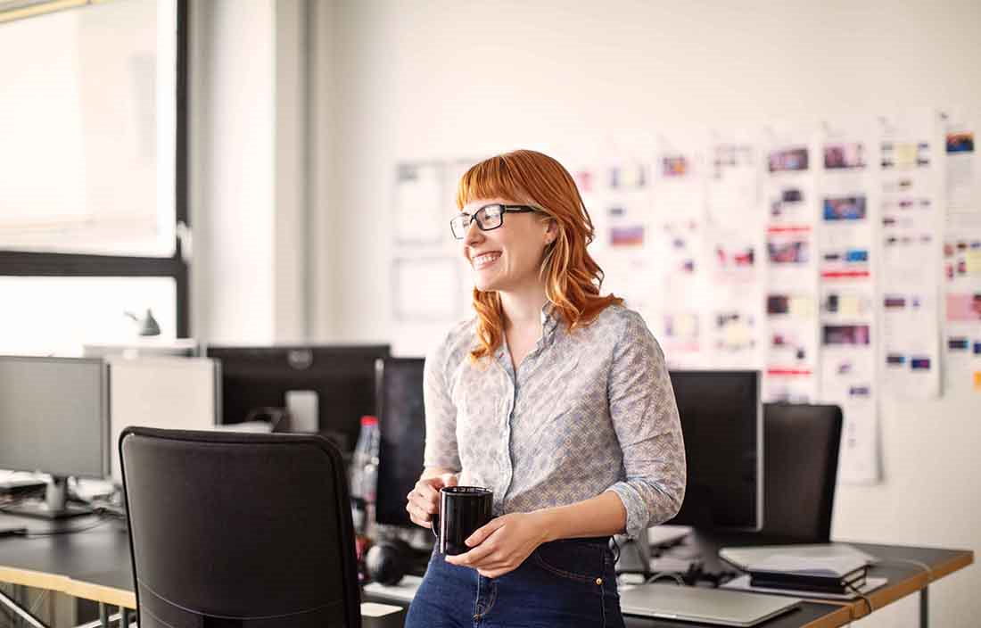 Red haired businesswoman smiling in the office holding a cup of coffee.