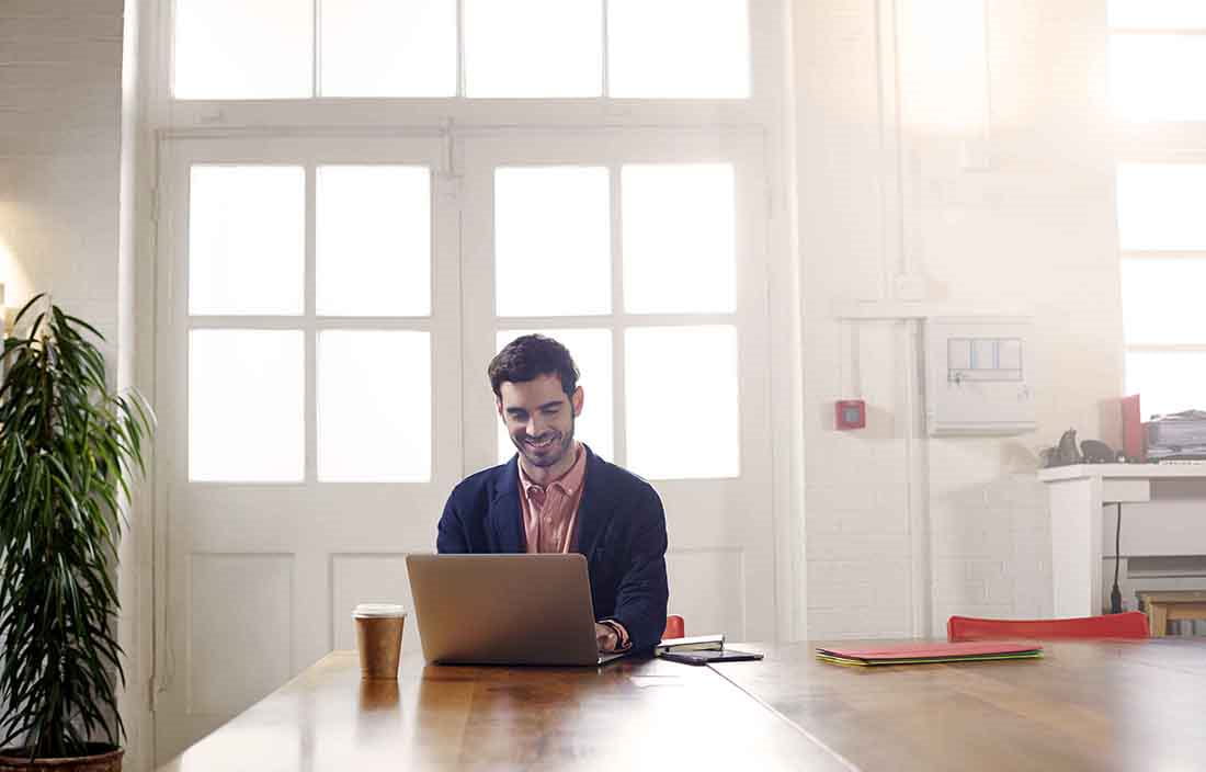 Businessman smiling while sitting at a kitchen table using a laptop computer.