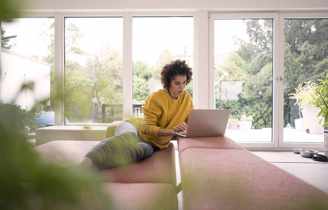 Woman leaning against a couch while using her laptop computer.