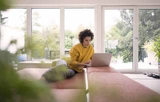 Woman leaning against a couch while using her laptop computer.
