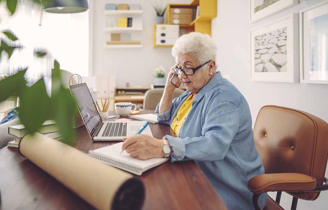 Elderly woman writing on a notepad at a desk.