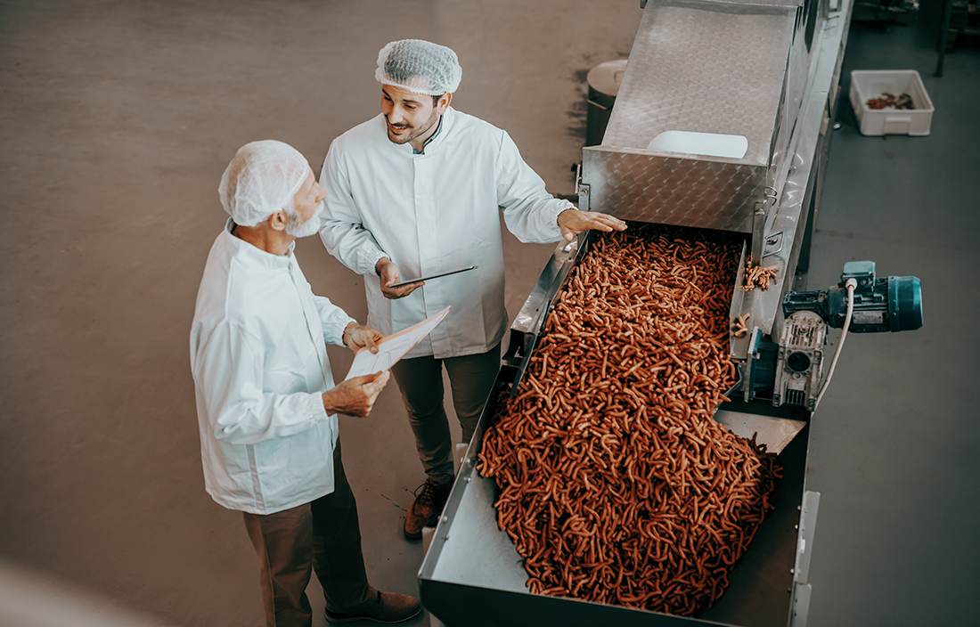 Two food workers in uniforms standing next to a conveyor belt.