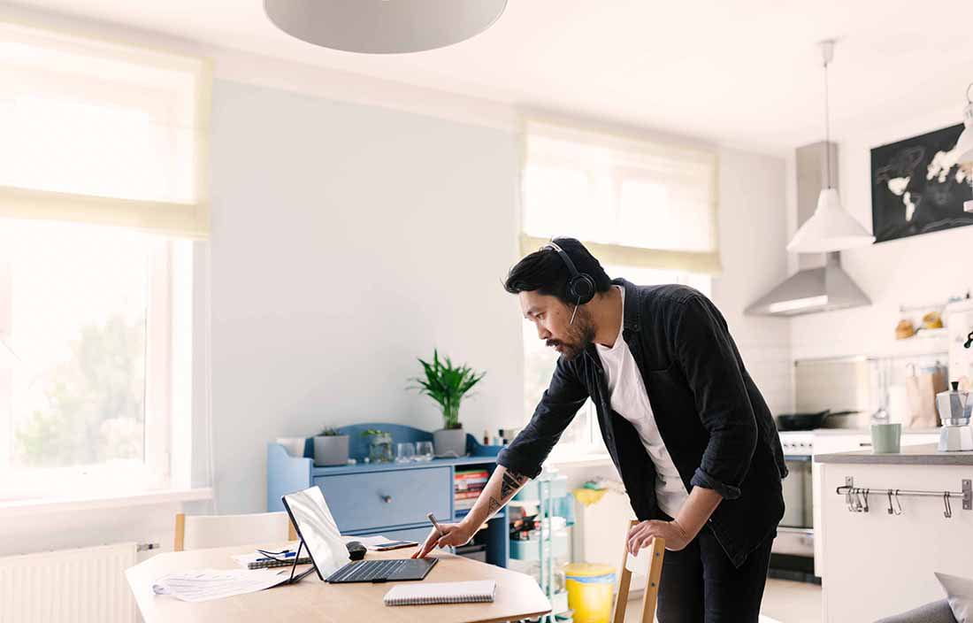 Man wearing headphones while leaning over a kitchen table using his laptop computer.