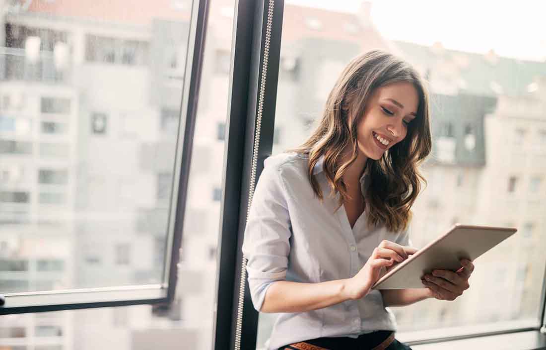 Business professional leaning against a window while using a tablet device.