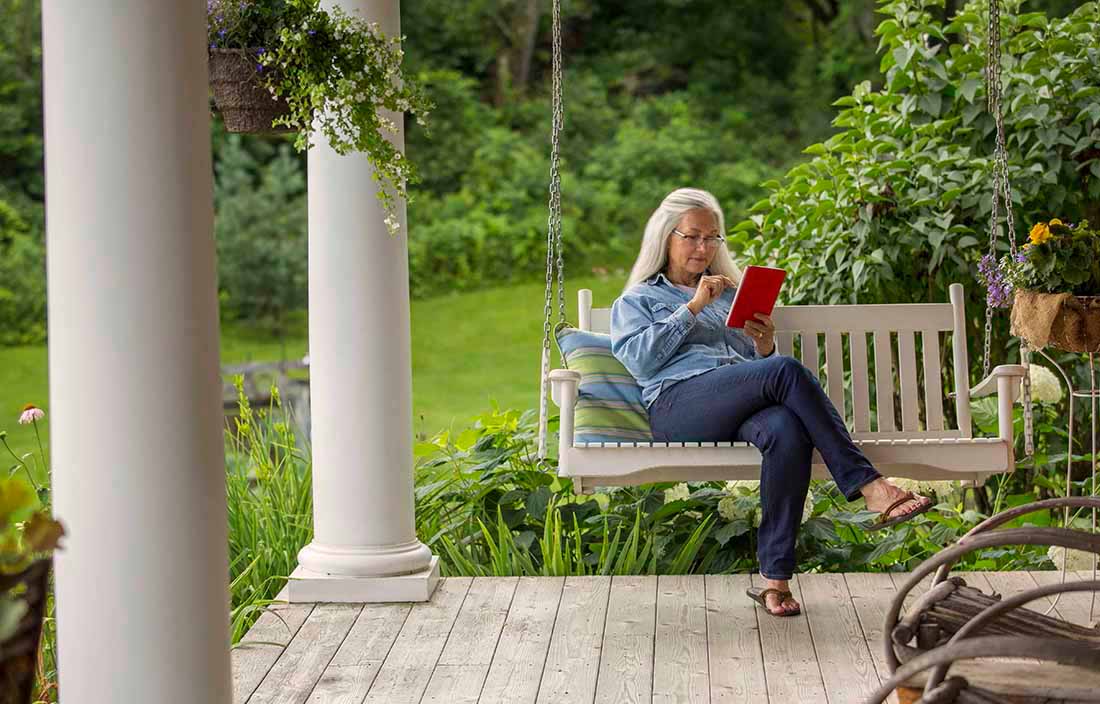 Elderly woman sitting on a swing on a porch using a handheld tablet device.