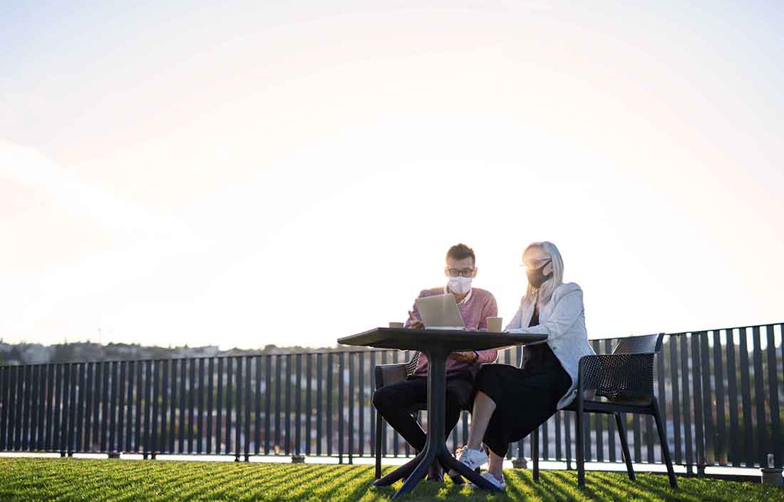 Two people wearing protective facemasks sitting at a table in a park.