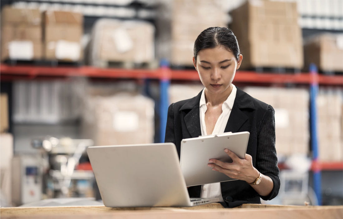 Business woman in a manufacturing warehouse checking inventory levels on a laptop computer.