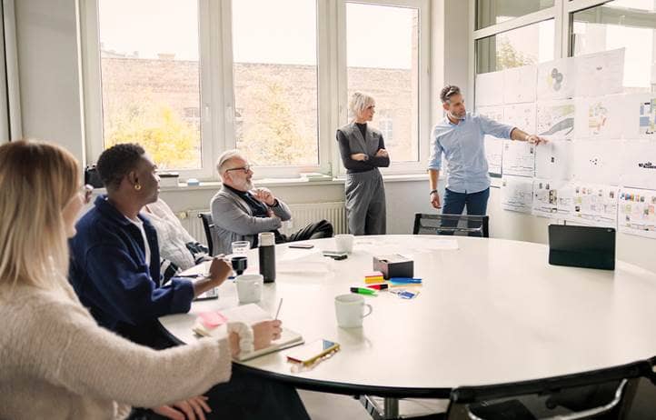 A group of people in an office watching a colleague present data on a whiteboard.
