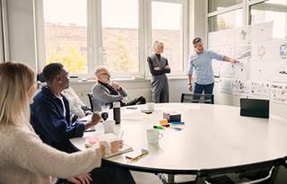A group of people in an office watching a colleague present data on a whiteboard.