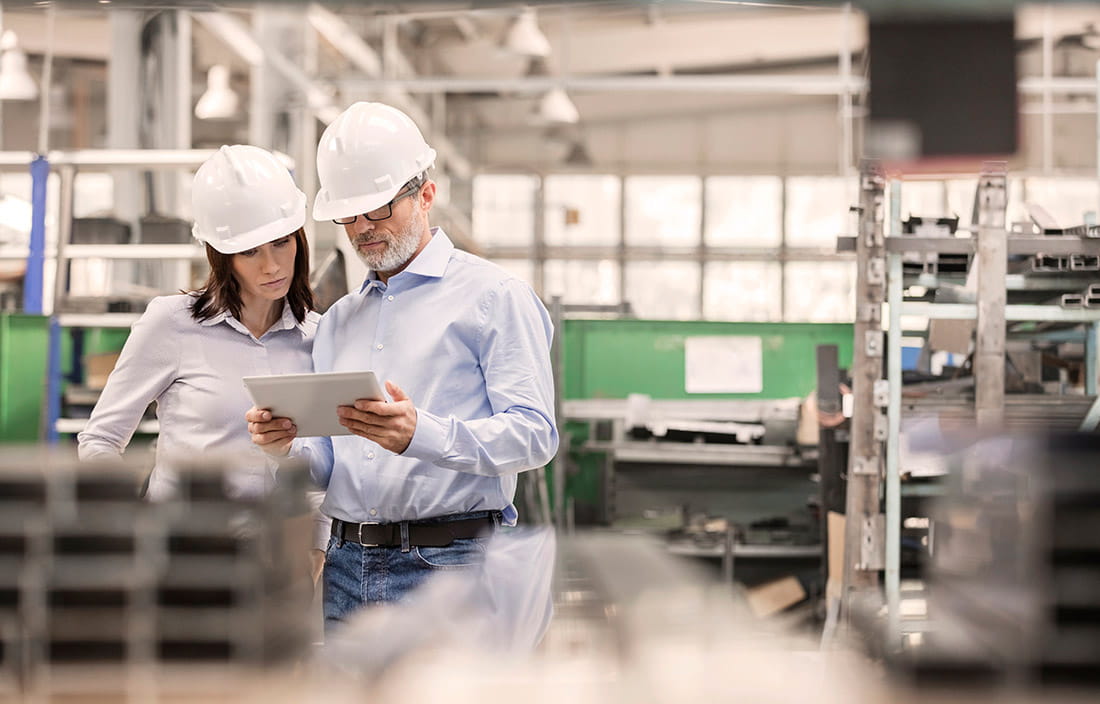 A man and woman standing in a factory wearing hard hats and reading an iPad